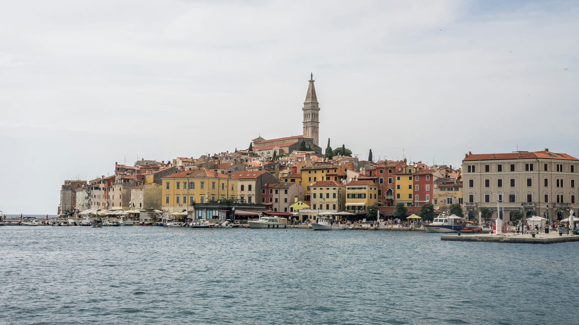 A quiet Adriatic waterfront under soft hazy light, with most of the café parasols folded away